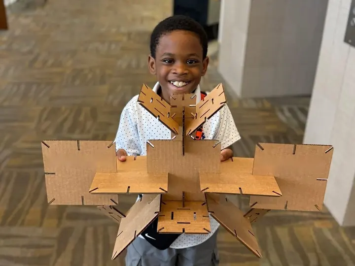 A smiling young boy holding up a large geometric sculpture made of laser-cut cardboard