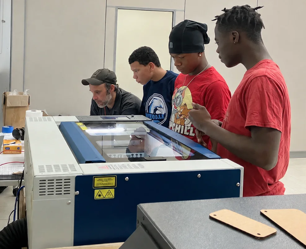 Sam Provenza at a laser cutter with three teens from Front Yard Bikes watching the cut