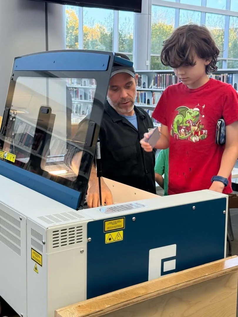 Sam Provenza at the laser cutter with a young maker, who is holding a piece just cut